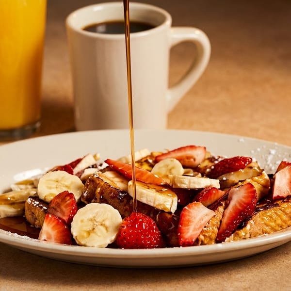 Syrup pouring over French toast with strawberries and bananas, with orange juice and coffee.