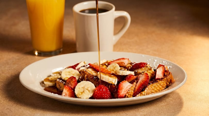Syrup pouring over French toast with strawberries and bananas, with orange juice and coffee.