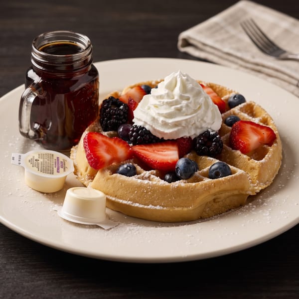 Golden waffle topped with whipped cream, strawberries, blueberries, and blackberries, with syrup.