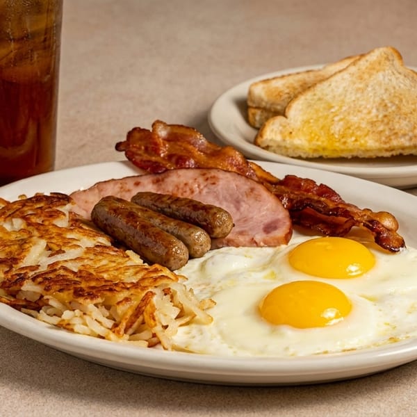 A hearty breakfast plate with fried eggs, hash browns, bacon, sausage, ham, toast, and iced tea.
