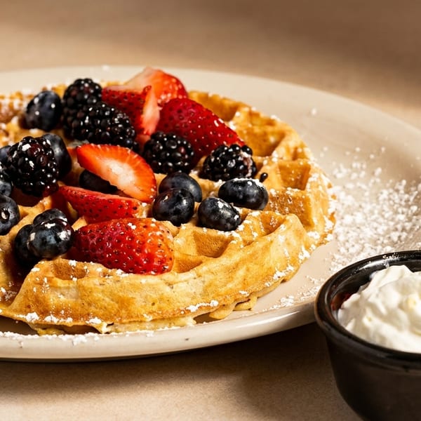 Golden waffle topped with fresh strawberries, blueberries, and blackberries, dusted with powdered sugar, with whipped cream.