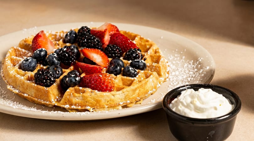 Golden waffle topped with fresh strawberries, blueberries, and blackberries, dusted with powdered sugar, with whipped cream.