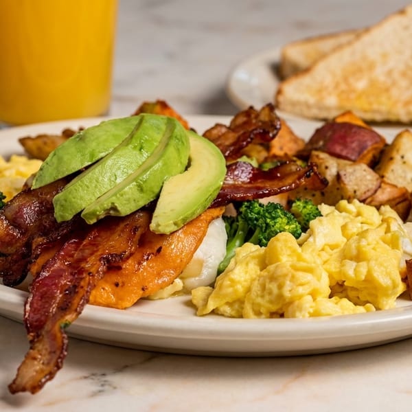 A breakfast plate with scrambled eggs, bacon, avocado, roasted potatoes, and broccoli.