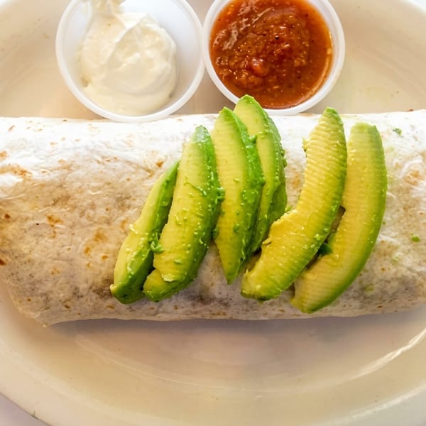 A large burrito topped with avocado slices, served with sides of sour cream and salsa.