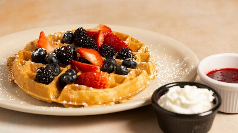 Golden waffle topped with fresh strawberries, blueberries, blackberries, powdered sugar, and sides of whipped cream and berry syrup.
